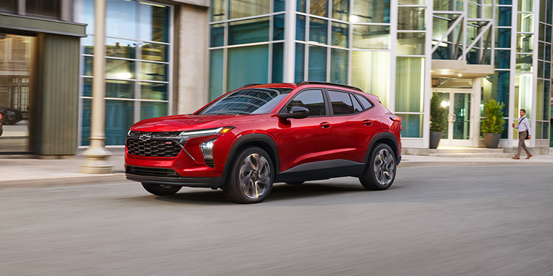 Red Chevrolet Trax driving through a city street, shown in a front three-quarter view with modern glass buildings in the background, highlighting its bold grille, sleek design, and sporty stance.