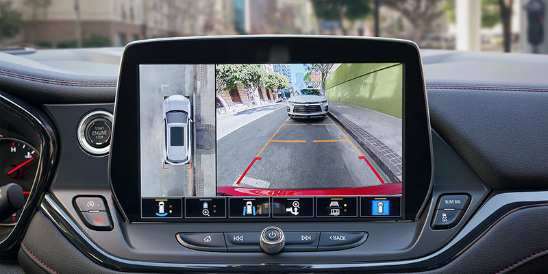Interior view of a vehicle showing a touchscreen display with a split-screen rearview camera system, including a top-down vehicle view and a rear camera view with parking guidelines.