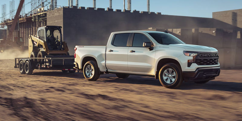 White Chevrolet Silverado pickup truck towing construction equipment on a trailer at a work site.